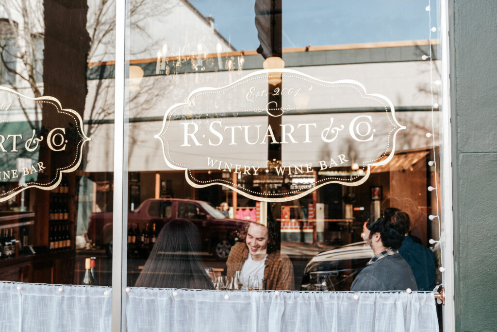 The exterior of a wine tasting room.  It has the logo of R. Stuart & Co. on a shiny glass window.  You can see three people seated inside tasting wine.