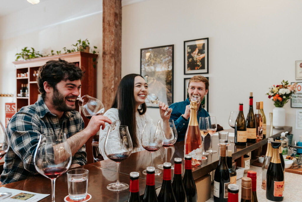 3 people sit at a bar.  They each have a few glasses in front of them with small amounts of wine.  They smile as they sip.