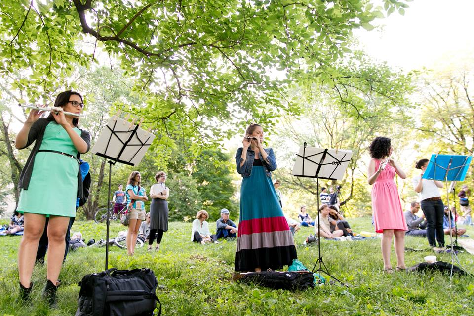 Three young women playing the flute stand in front of music stands, while a crowd watches in a grassy field.