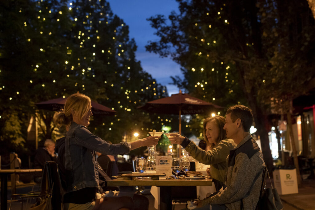 A table full of people sit at night, lit by twinkle lights in the trees and street lamps.  Two women cheers.