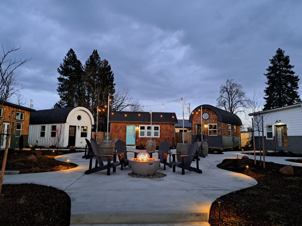 It's dusk, and a courtyard is lit with gold lights and a fire pit.  5 tiny houses are in the background.