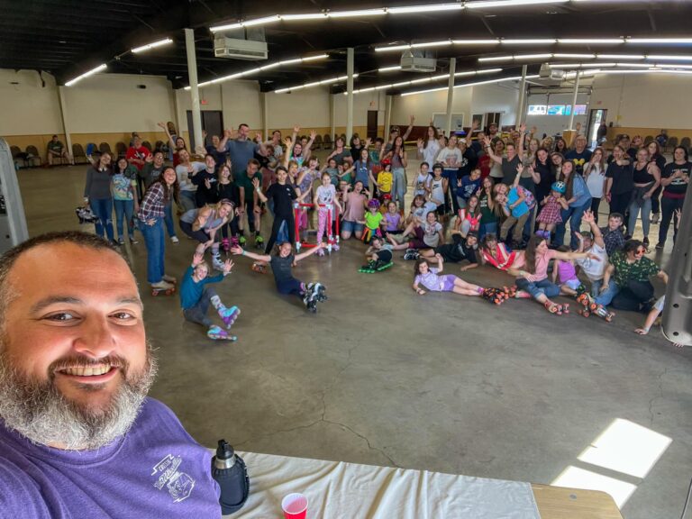 A man takes a selfie with a large group of people who are all wearing skates and posing for a group shot in a large indoor space.