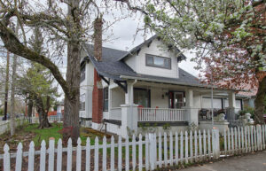 A white craftsman style bungalow with black trim. It has a white picket fence around the yard.