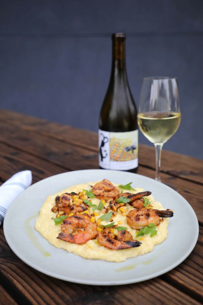 A white plate with 6 grilled prawns served atop a generous bed of polenta.  It is garnished with roasted corn and parsley.  Behind it are a glass of white wine and a wine bottle.