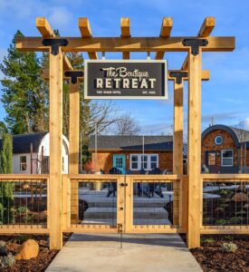 A wood and metal gate holds a sign that reads 'The Boutique Retreat'. Behind it, you can see three tiny houses and a courtyard.