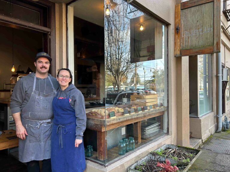A smiling couple with aprons on stand with their arms around each others' backs. A restaurant is behind them with a glass window. You can see an array of baked goods.
