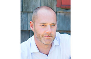 A headshot of a man with very short hair and a five o'clock shadow