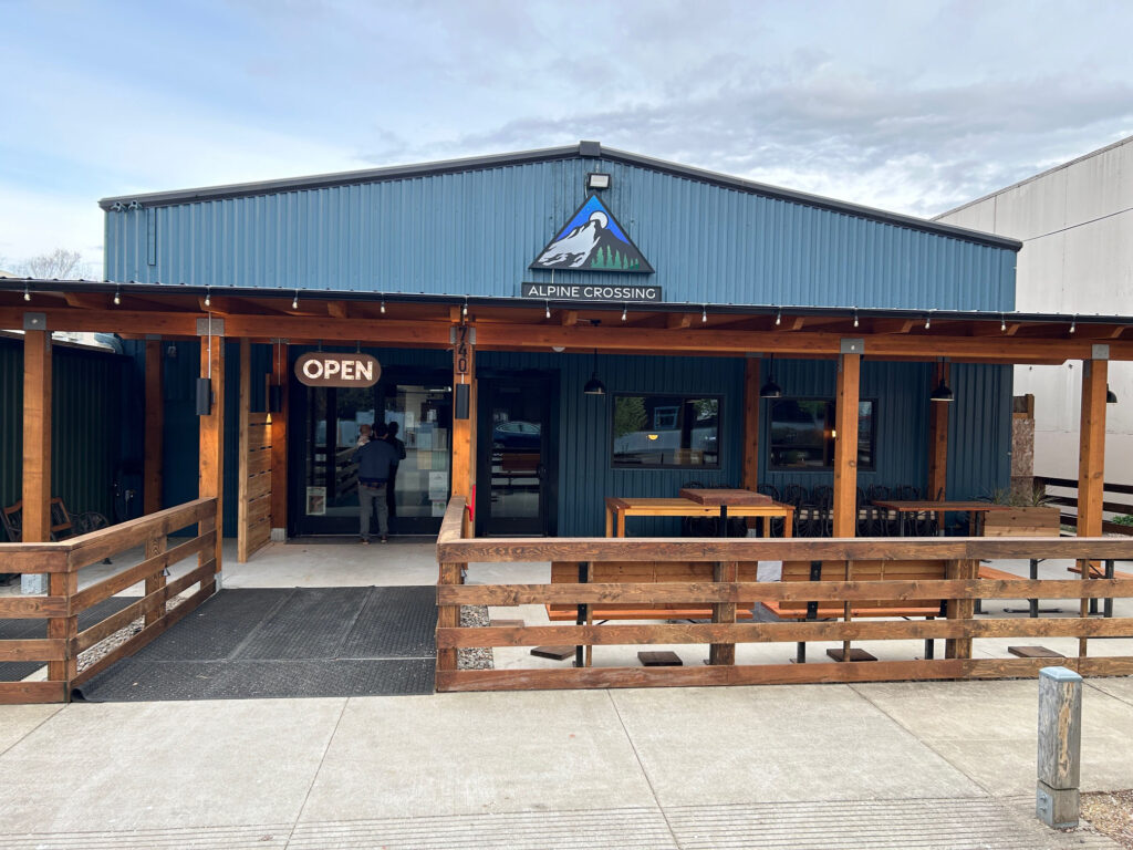 The exterior of Alpine Crossing.  It is a blue building with corrugated metal siding.  There is a covered patio and tables and chairs.  Someone walks into the door underneath an open sign.