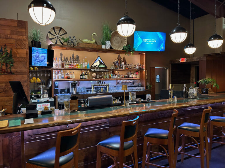 A shiny wood bar with backed barstools. There are two tvs and a fully stocked bar.