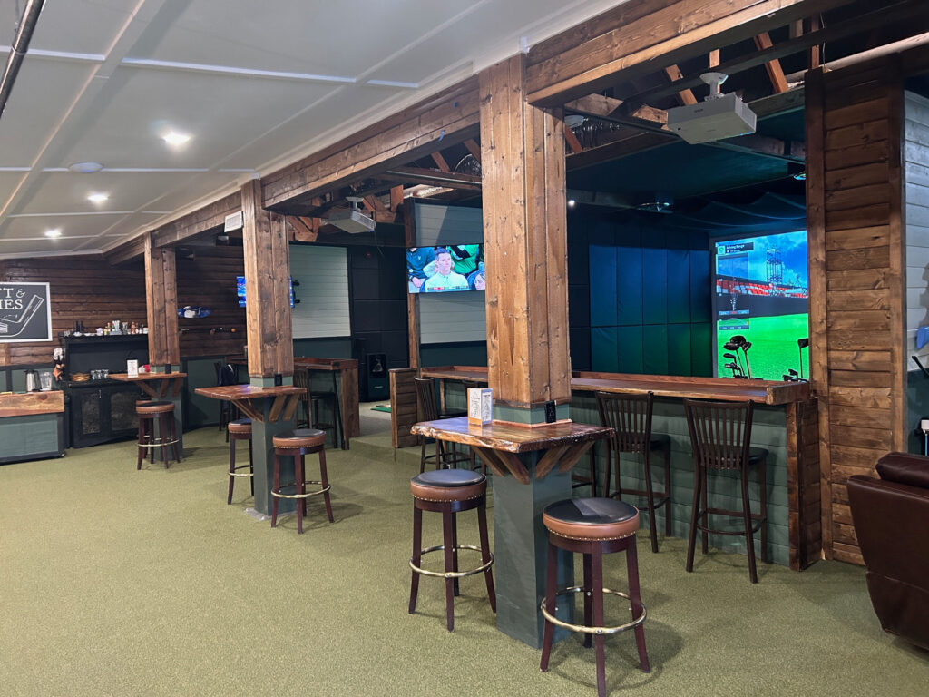 Stools are pushed up against wood tables and bars.  There is green carpet on the floor.  There is simulated golf in a nearby nook.