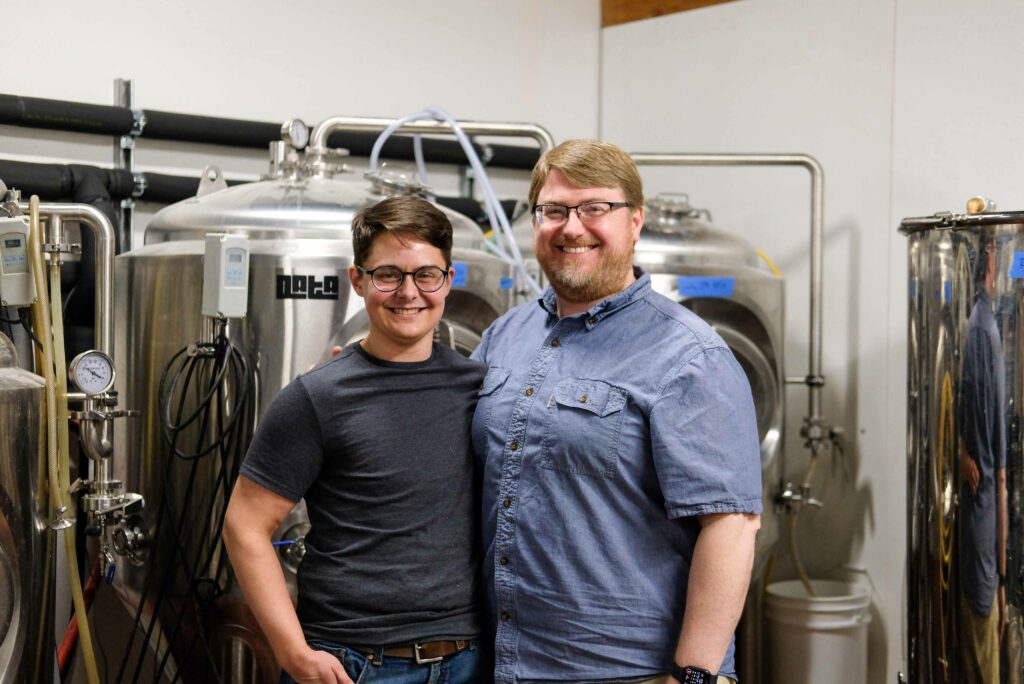 Two smiling men stand next to each other, with their arms around eachothers' shoulders.  They are in front of beer tanks.