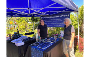 Two women stand and smile under a blue canopy. A sign behind them says The Carlton Observatory Project.