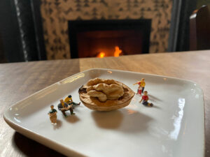 The Tiny Travels figurines sit atop a white plate enjoying the view of a cracked walnut and a fire in the background.