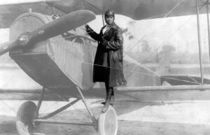 A back woman stands on the tire of a plane. She is wearing aviation gear.