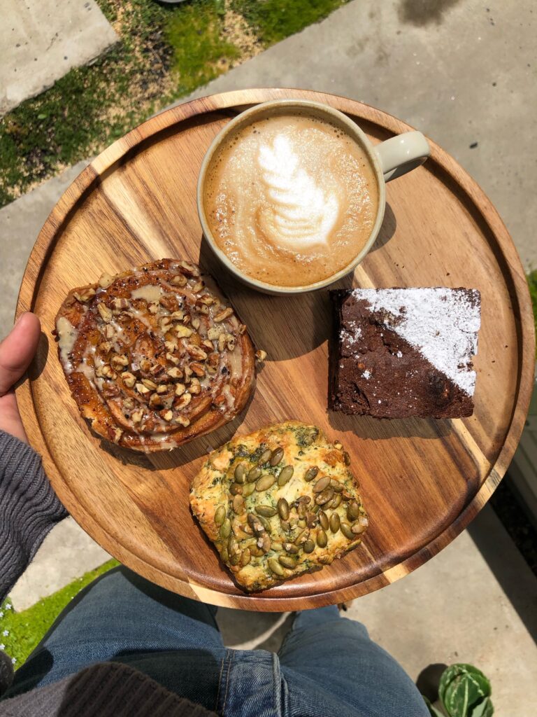 A latte, brownie, pecan roll and toast with pumpkin seeds sit atop a wooden tray.