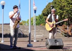 One man plays a saxophone and another plays the guitar while singing.