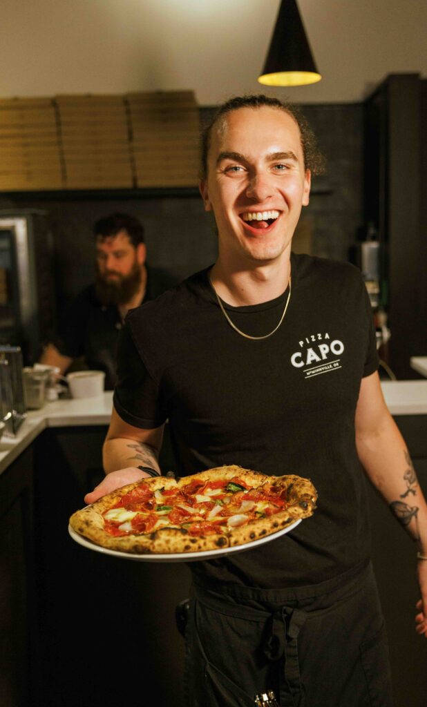 A young man with a large grin on his face and a t-shirt that says Pizza Capo holds a pizza.  In the background, a bearded man prepares food in the open kitchen.