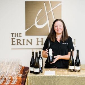 A woman holds a botthle of Chris James Cellars wine. The logo for The Erin Hanson Gallery is behind her.