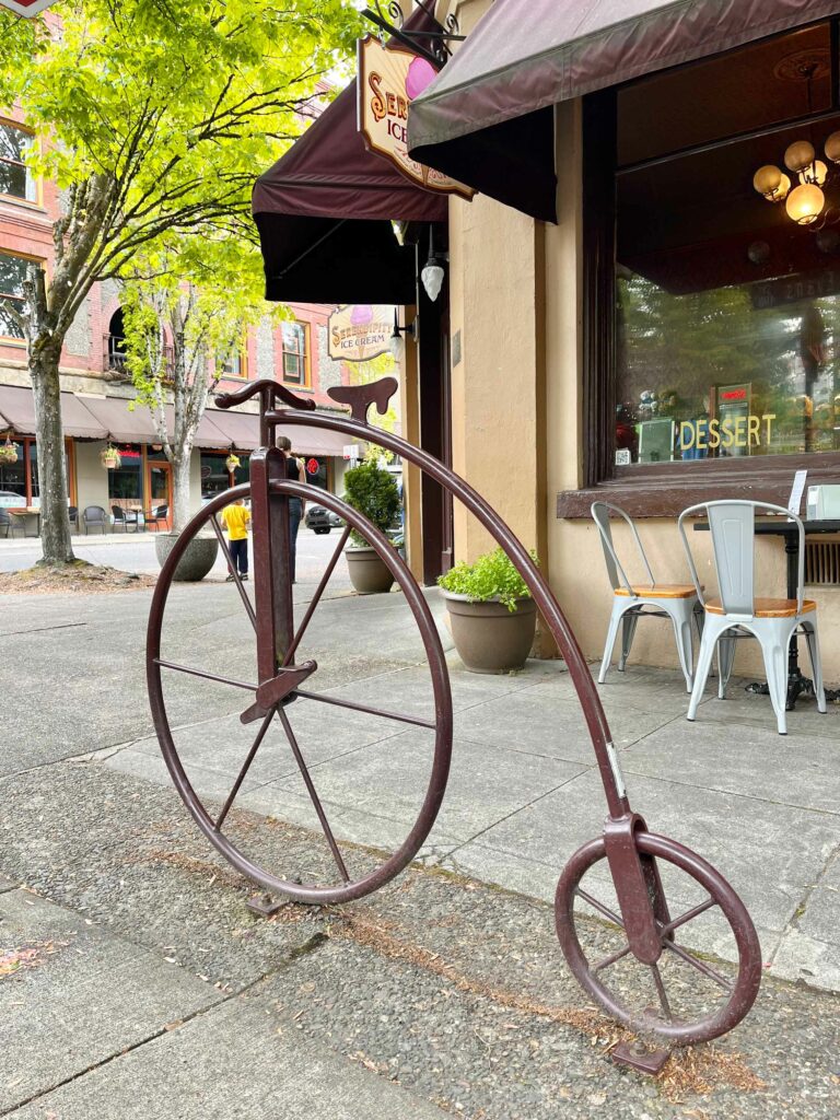A metal bike rack shaped as a penny farthing bike with Serendipity Ice Cream shop in the background.