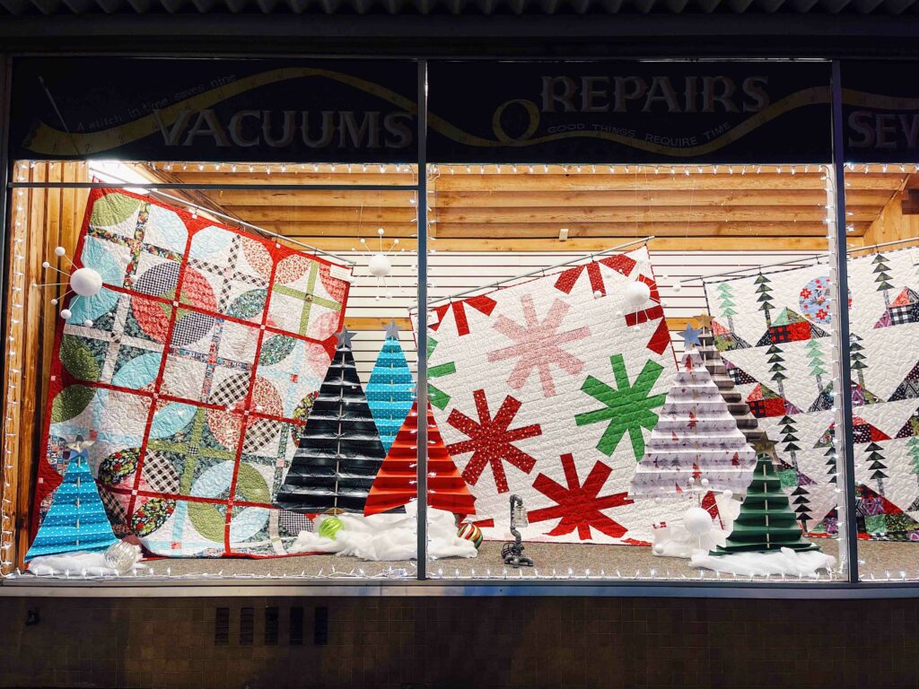 A retail window display of holiday quilts are hung and displayed along with paper Christmas trees and cotton snow.