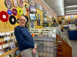 A man stands in front of a display of cds, a wall of records and his record store in the background.