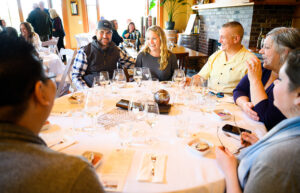 A group of people sit around a round table. They all have glasses of wine.