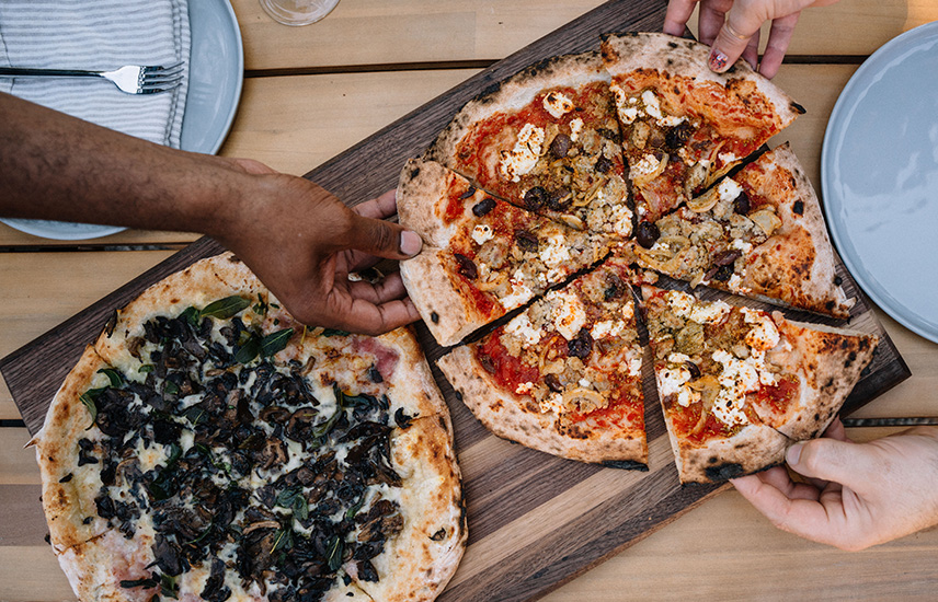 Two pizzas are on a cutting board.  Three people each grab a slice.