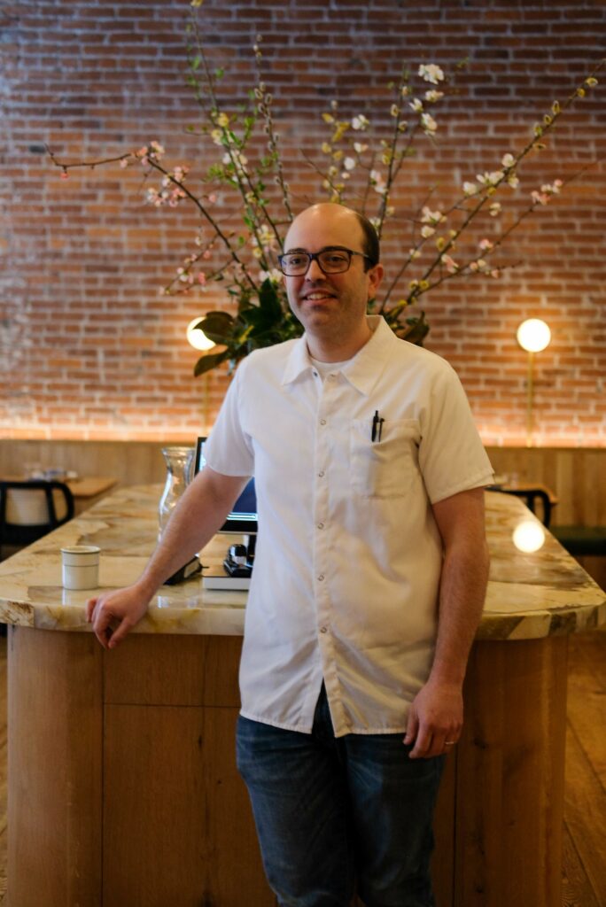 A smiling man with glasses stands in front of a marble countertop and a flowering plant behind him.
