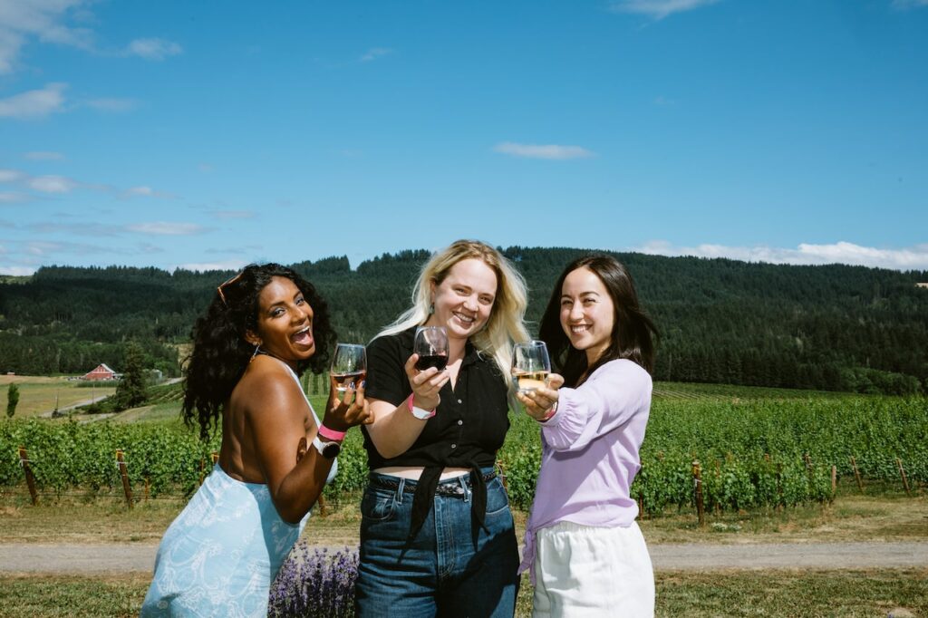 Three smiling women hold up stemless wine glasses with different colored wine in them.  Vineyards and tree lined hills are in the background.