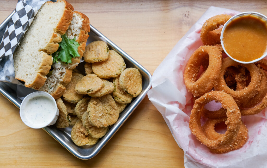 A metal tray with a hearty looking chicken salad sandwich with coins of breaded and fried pickles and a paper ramekin with ranch.  There is also a paper lined red basket with onion rings and a large paper ramekin with orange dipping sauce.