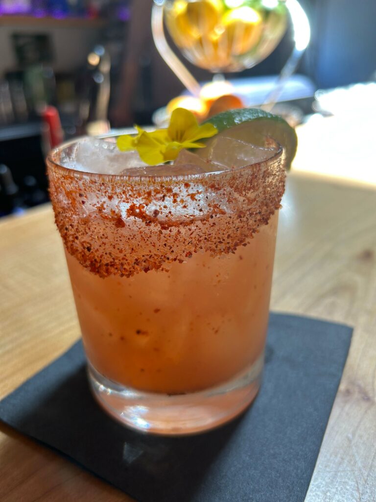 A rocks glass is filled with a pinkish red liquid and ice.  The rim is sprinkled with a red powder.  A yellow pansy and a slice of lime are garnishes on top of the drink.