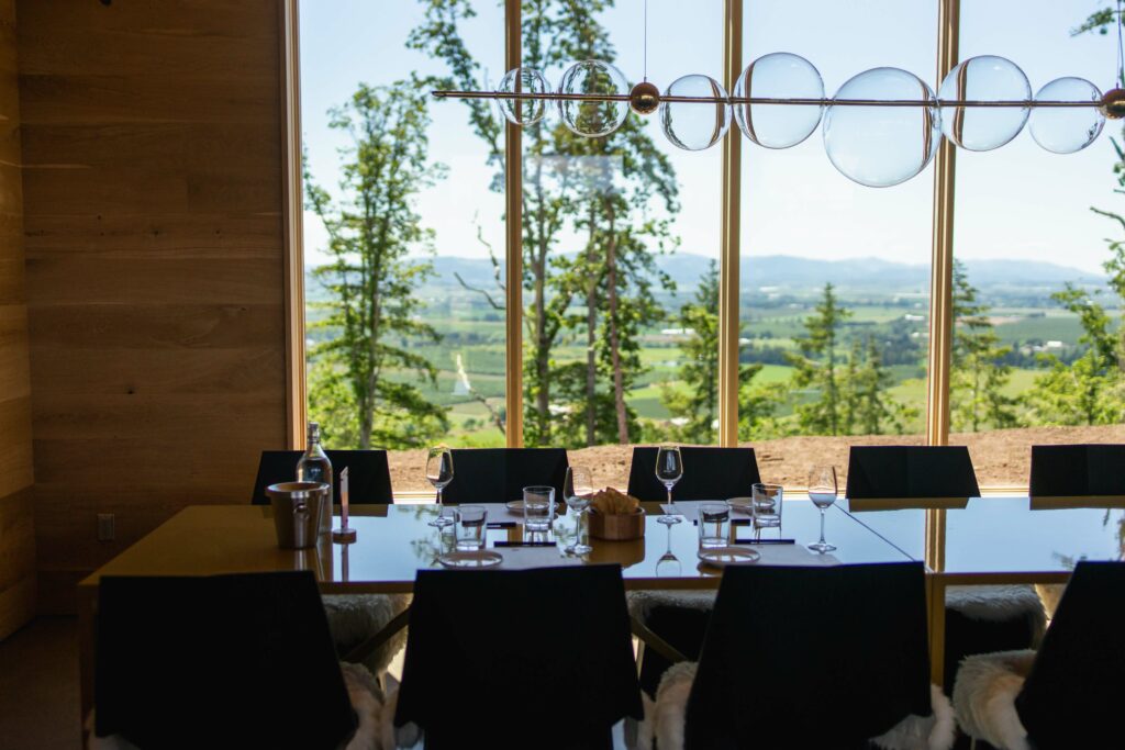 A long table is set up with wine and water glasses and small plates.  A light fixture that looks like bubbles is right above it.  The table is in front of a large window that overlooks a sprawling view of lush trees and greenery.  The hills are in the distance.