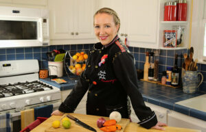 A woman in a chef's uniform stands in a kitchen in from of a counter with a cutting board with peppers, onions, limes and a knife.