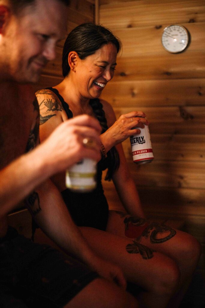 Two people smile and lift cans of Bierly Brewing beer up to their mouths.  They are in a wood lined sauna.