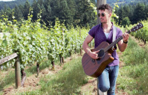 A man with a guitar stands in a vineyard. He is leaning against a post and strums.