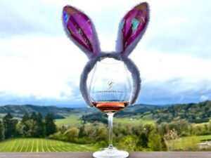 A glass of rose wine is wearing a bunny ear headband. A vineyard is in the background.