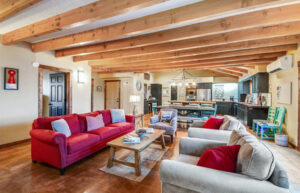 A large family room with wood beams spanning the ceiling. There's a red sofa, two white overstuffed chairs and a coffee table. A dining table is in the background.