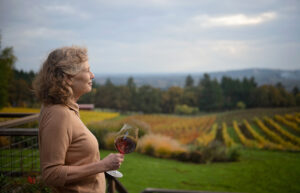 A woman smiles, holding a glass of read wine, as she looks out across a vineyard.