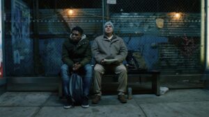 Two men sit on a bench in front of a chain link fence with industrial looking lights strung across it.