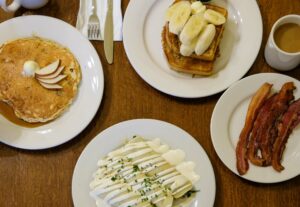 A spread of breakfast dishes, including pancakes, french toast with sliced bananas, crepes with a white sauce drizzled on top, a side of bacon and a cup of coffee.