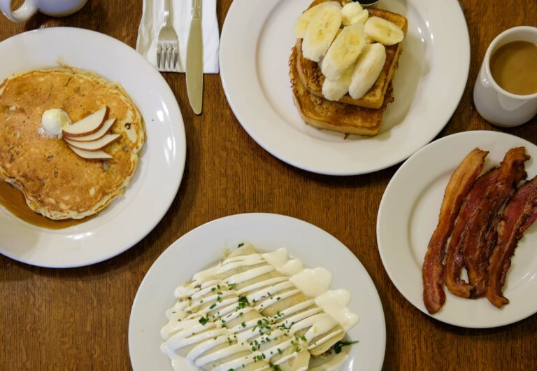 A spread of breakfast dishes, including pancakes, french toast with sliced bananas, crepes with a white sauce drizzled on top, a side of bacon and a cup of coffee.