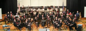 A large group of band members sit in concert formation with their instruments.