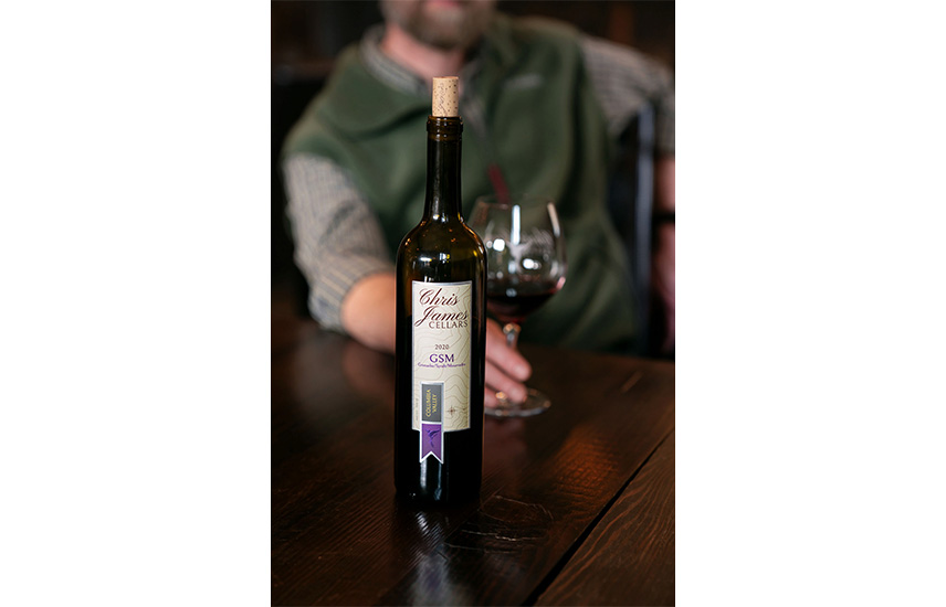 A bottle of Chris James Cellars is on the table. A person pushes a wine glass with red wine forward.