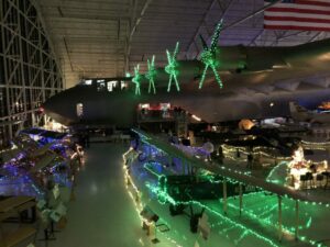 The Spruce Goose airplane is inside of a large museum. Its propellors are decorated with green Christmas lights. A biplane is next to it and is also decorated with green Christmas lights.