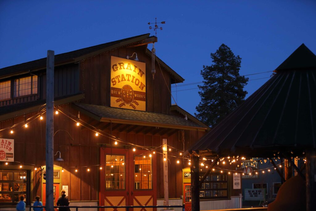 It is nightfall.  A large barn has a sign that says Grain Station Brew Works.  There is string cafe lighting strung around an outdoor courtyard.