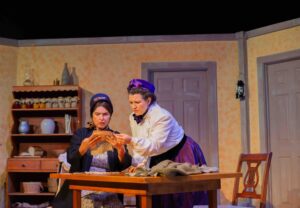 Two women in period costume lean over a table, looking at a piece of fabric, with concerned looks on their faces.