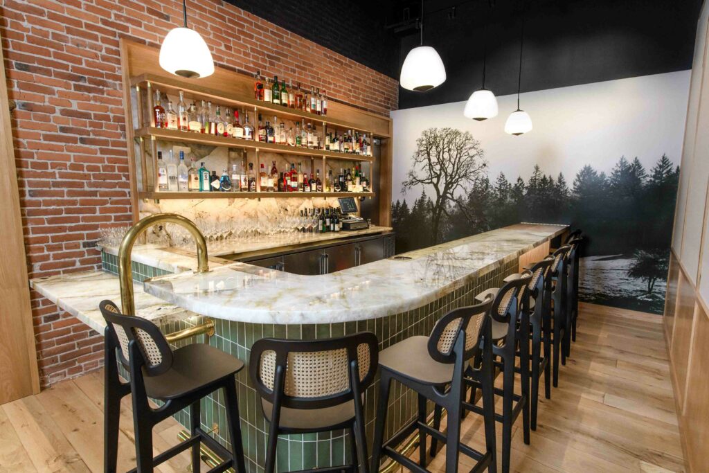 A large quartzite bar with green tile below.  Chairs span the bar and pendant lighting hangs over it.