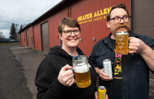 Two people stand in front of Heater Allen brewery. They smile and hold large beer mugs.