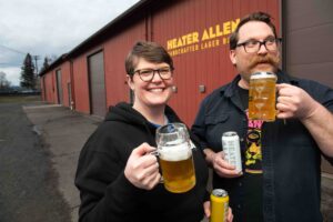 A man and a woman hold full beer steins in one hand and a can of Heater Allen beer in another. Behind them is a warehouse that says Heater Allen.