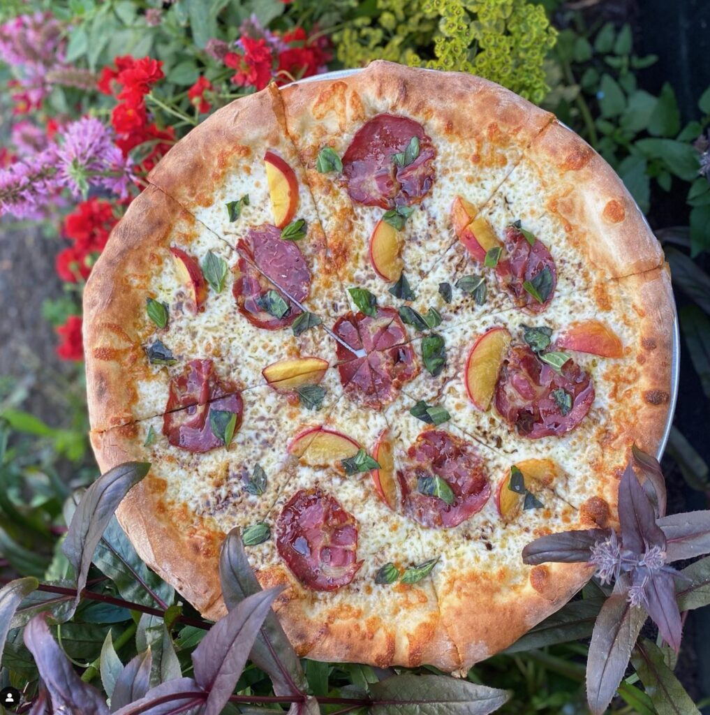 A big beautiful pizza with peaches, basil, meat and cheese.  The crust is cooked perfectly to a golden brown.